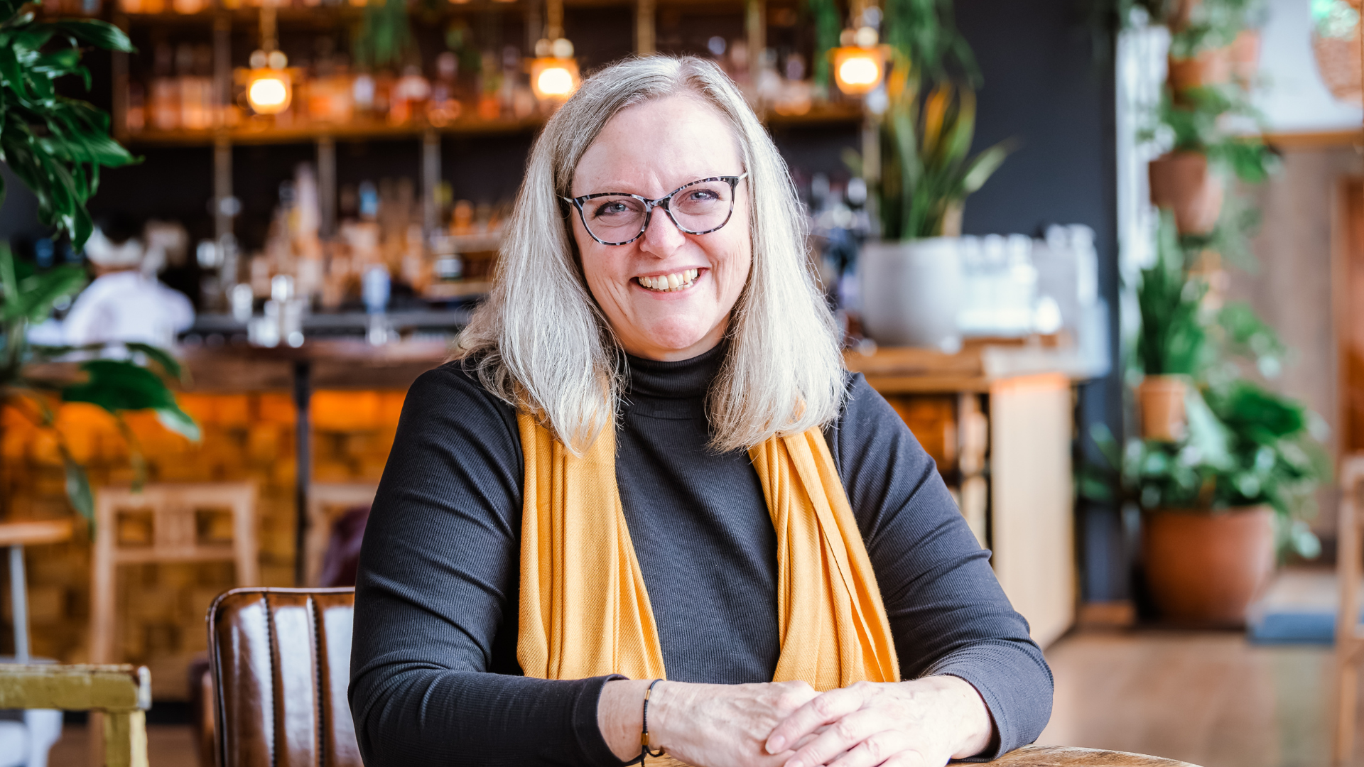 A smiling woman, Annie Bennett, with shoulder-length grey hair and glasses sits at a wooden table in a cozy, warmly lit café. She wears a black top and a mustard yellow scarf, with her hands resting on the table. The background features a stylish bar area with hanging lights, plants, and shelves of glassware.