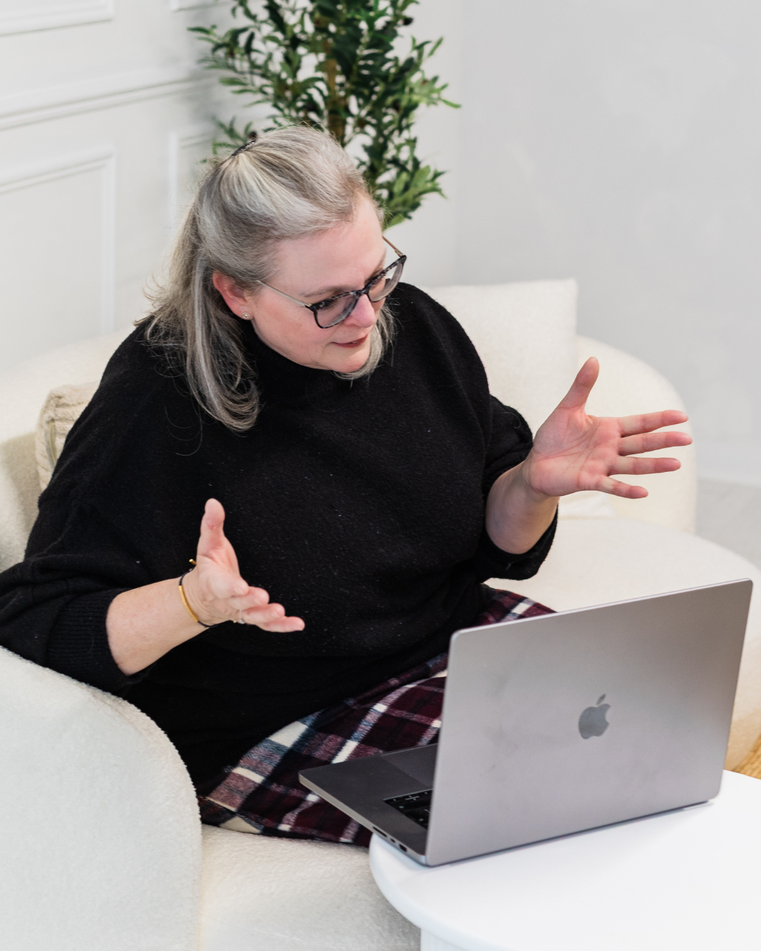 A woman with grey hair and glasses sits on a cream armchair, gesturing as she talks to a laptop during an online meeting.
