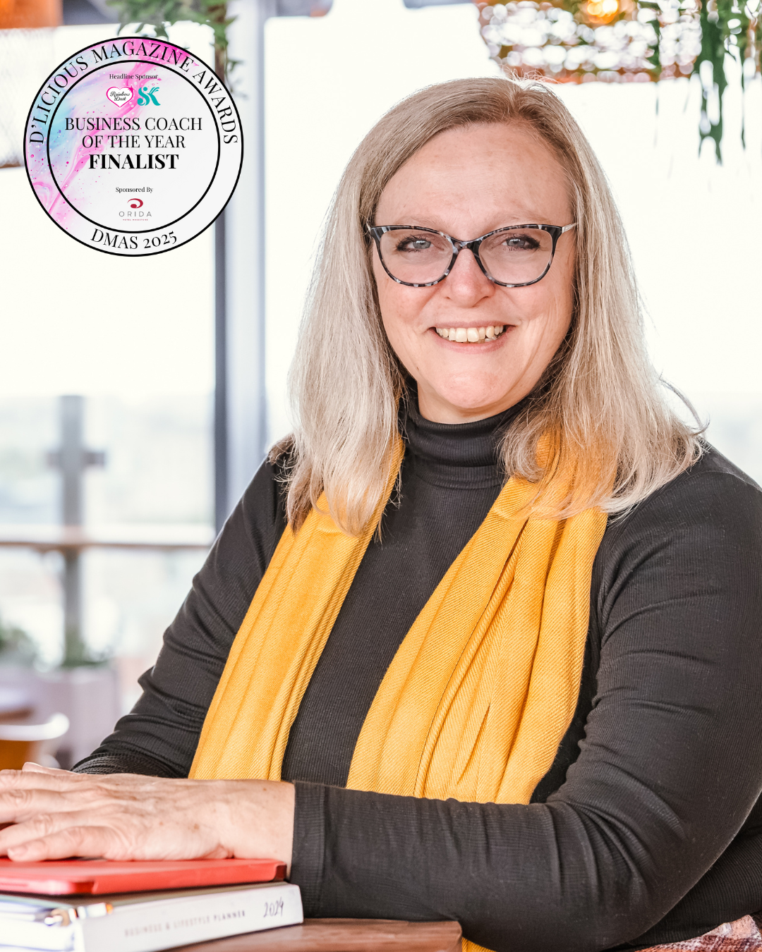 Professional portrait of Annie Bennett, business coach and mentor, smiling while seated at a table in a light, modern space. She wears glasses, a black top and a mustard scarf, with a Delicious Magazine Awards – Business Coach of the Year Finalist 2025 emblem visible.