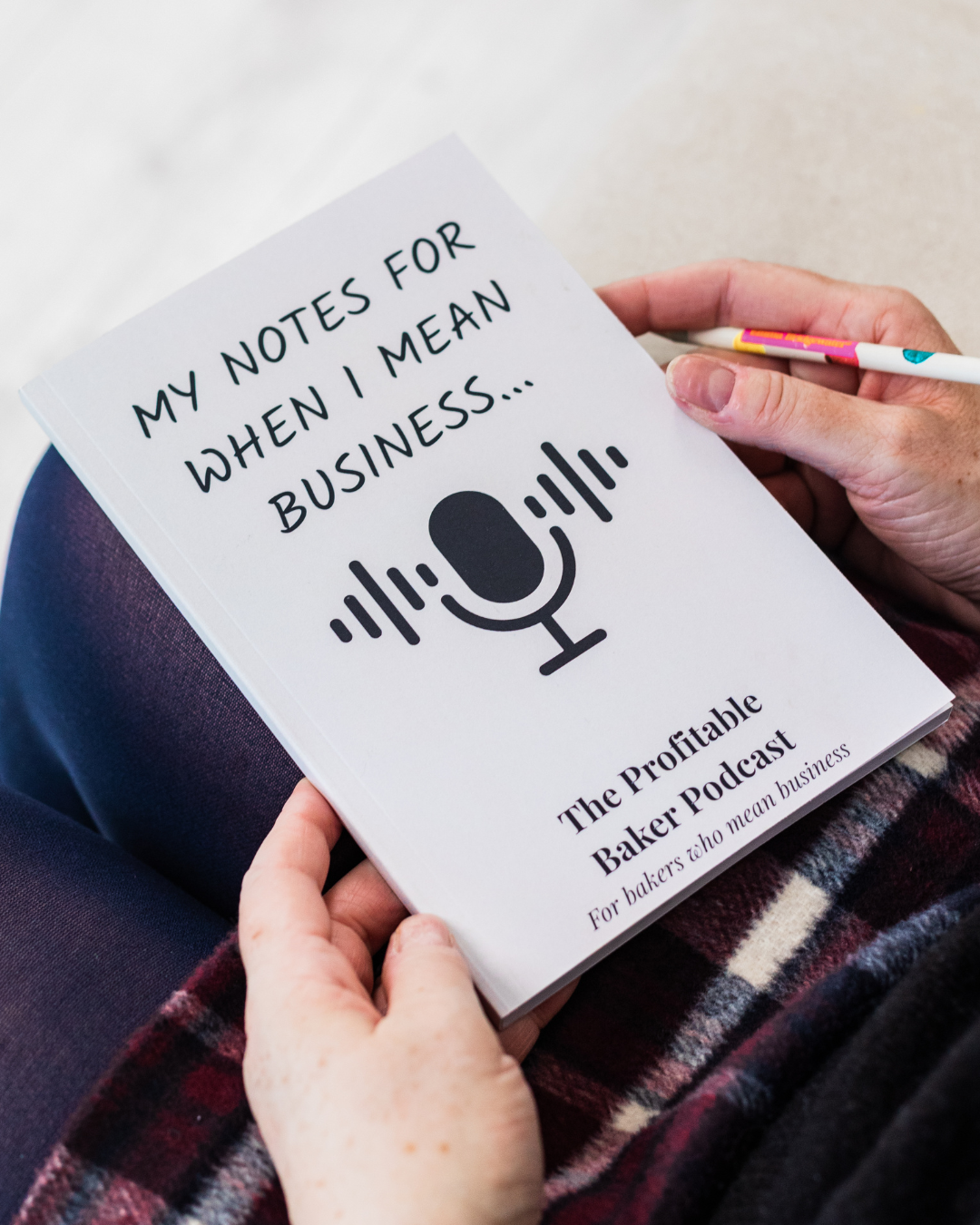 A close-up of hands holding a white notebook titled "My Notes for When I Mean Business..." featuring a podcast microphone graphic, branded for The Profitable Baker Podcast — "For bakers who mean business."
