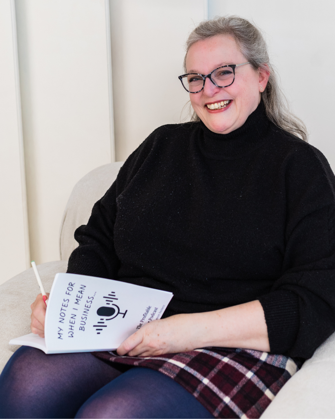 A smiling woman with grey hair and tortoiseshell glasses, wearing a black turtleneck sweater and plaid skirt, sitting in a cream armchair holding The Profitable Baker Podcast branded notebook and a pen.
