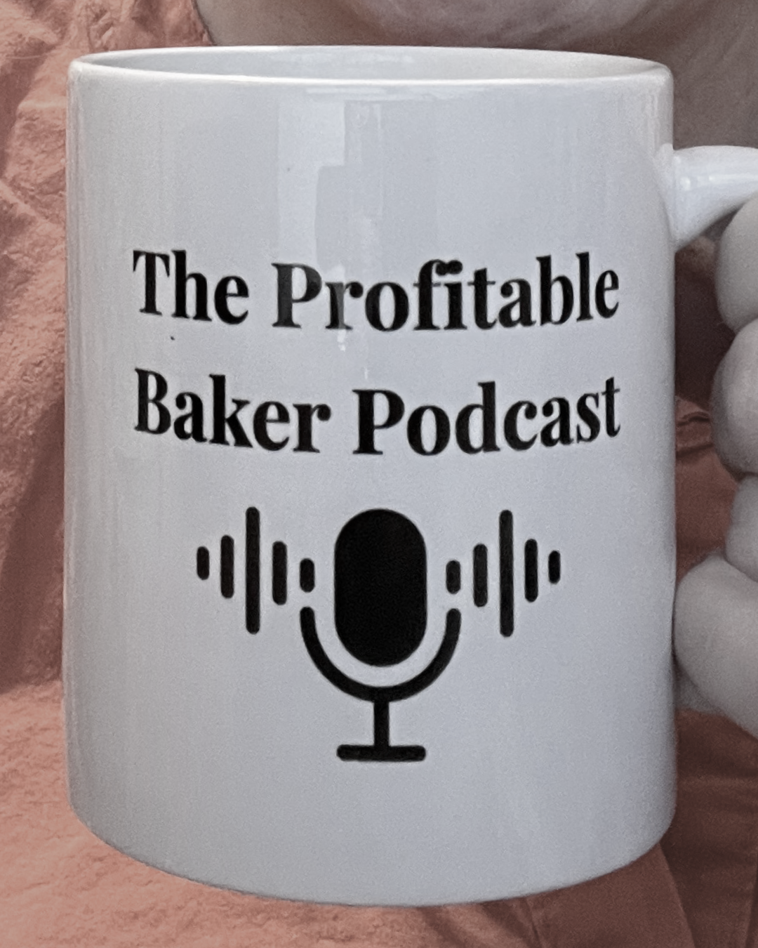 A white ceramic mug displaying "The Profitable Baker Podcast" in bold serif text above a podcast microphone with sound wave graphics, set against a pink background.