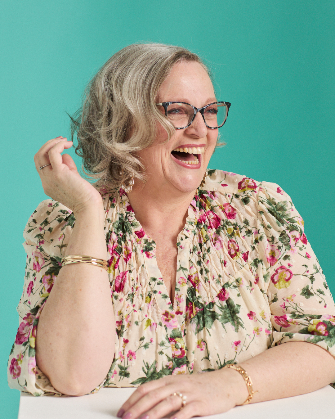 Annie Bennett laughing and looking to the side, wearing a floral blouse and gold jewellery, against a bright teal background.