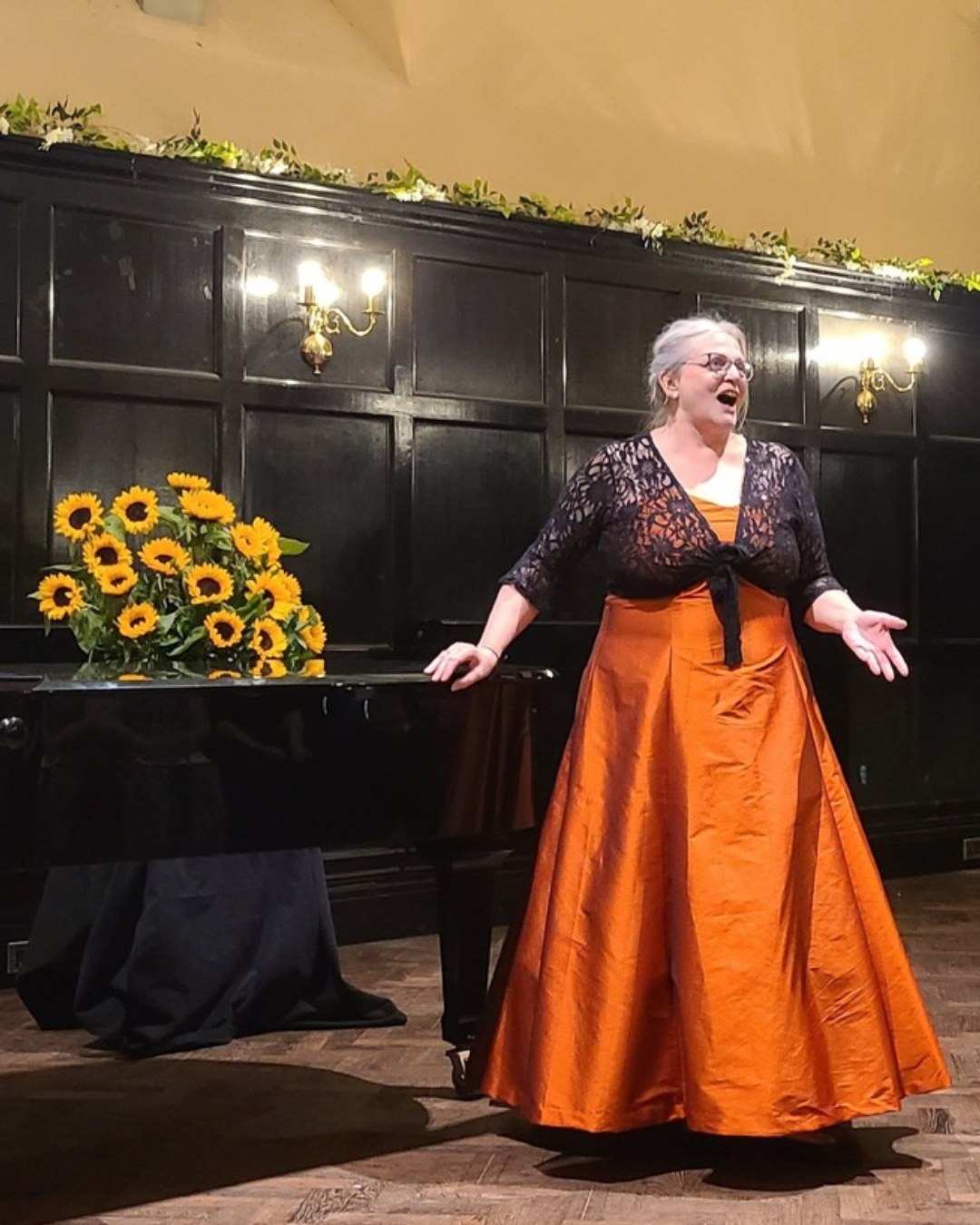 Annie Bennett performing on stage in a sweeping orange silk skirt and black lace top, arms open wide, with a grand piano and large bouquet of sunflowers behind her in an elegant panelled room.