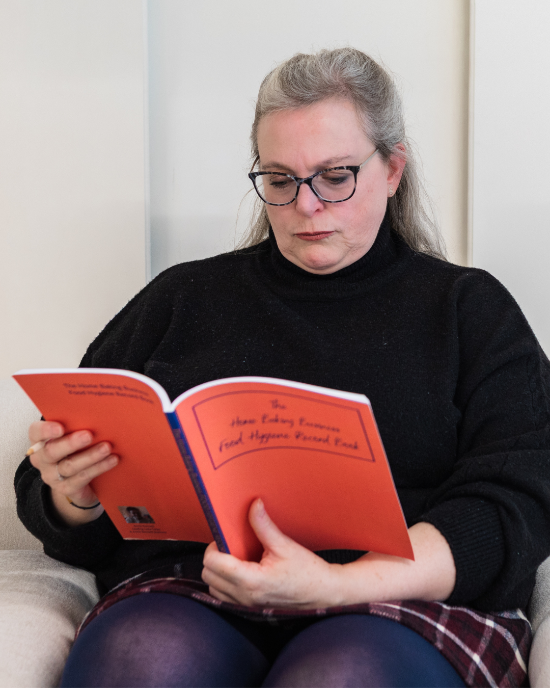 Annie Bennett sitting in a chair reading her book, The Home Baking Business Food Hygiene Record Book, which has a bold orange cover.