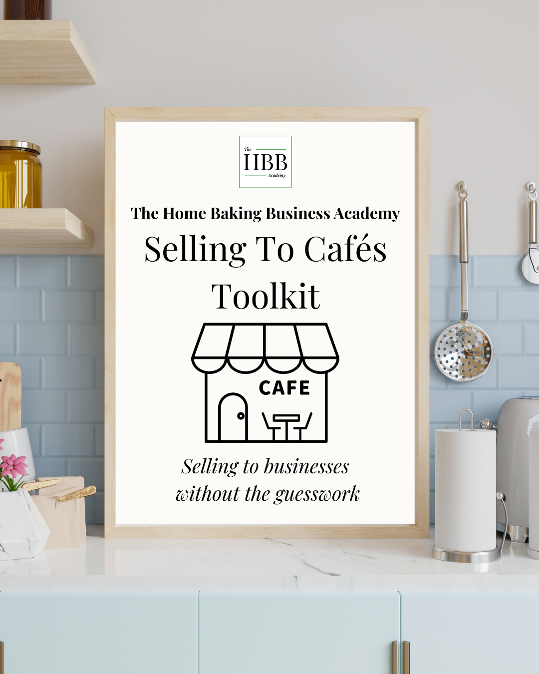 Promotional graphic for the Home Baking Business Academy's Selling To Cafés Toolkit, taglined 'Selling to businesses without the guesswork', displayed as a framed poster on a kitchen counter