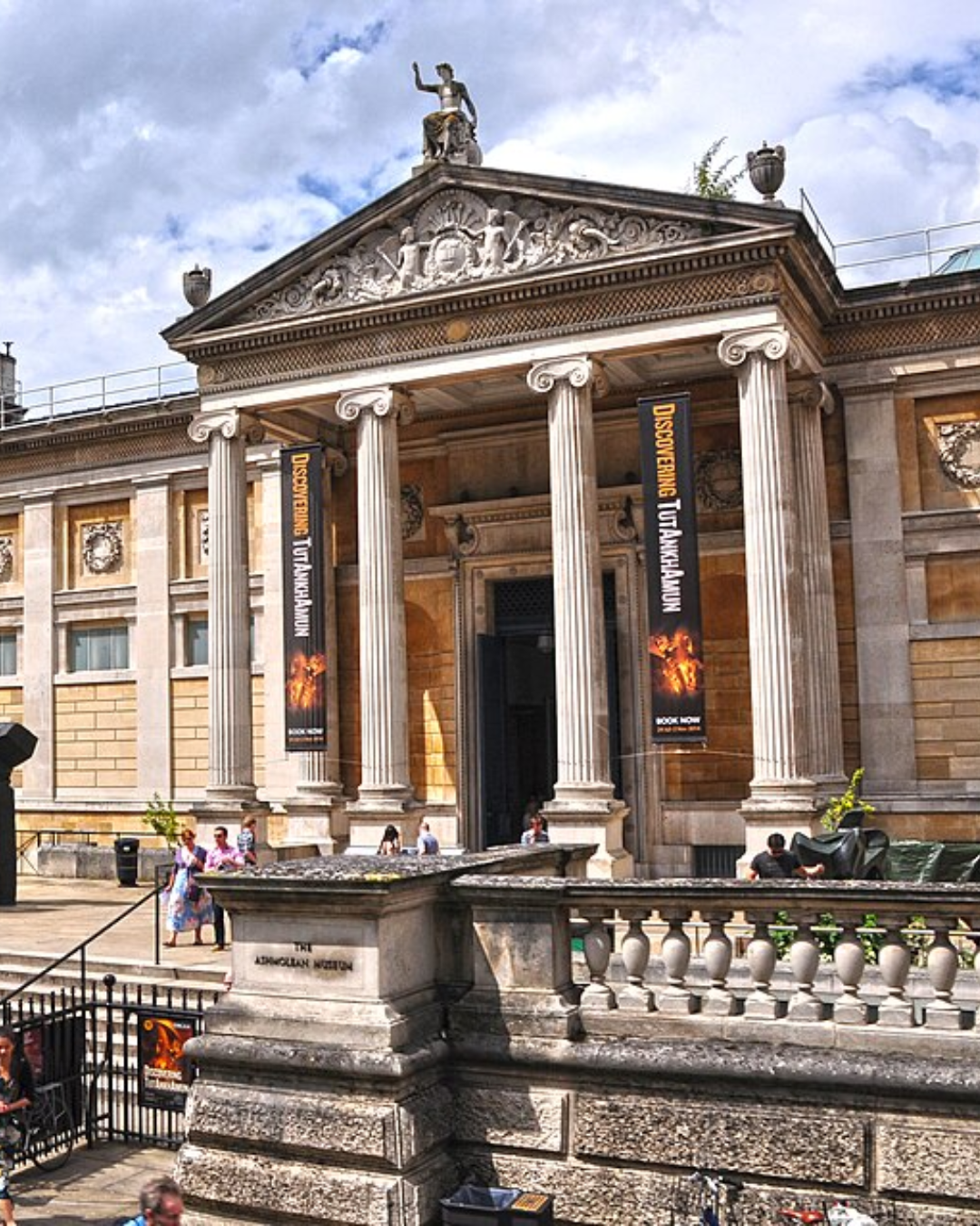 The neoclassical entrance to the Ashmolean Museum on Beaumont Street, Oxford. Stone columns frame the grand portico, with promotional banners for a Tutankhamun exhibition hanging between them. Visitors sit on the steps in summer sunshine.