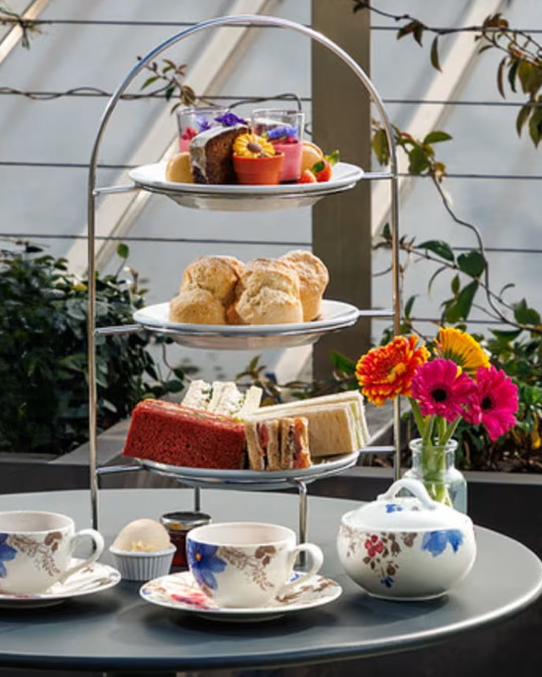 A three-tiered afternoon tea stand on a grey table, set against a glass conservatory backdrop with greenery. The stand holds finger sandwiches and a slice of red velvet cake on the lower tier, scones on the middle tier, and decorated cakes and sweet treats on the top tier. Floral teacups and a teapot sit alongside a small vase of bright orange, pink and yellow gerberas.