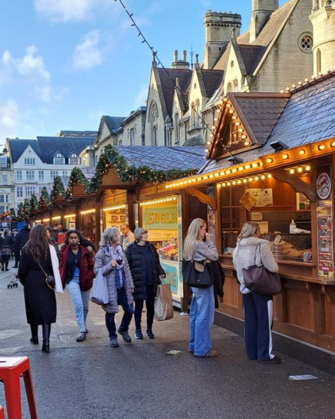 A row of wooden Christmas market chalets lit with warm fairy lights and decorated with garland, photographed at night against a honey-coloured stone building. Two visitors in winter coats browse the stalls. A colourful wristband display is visible on the right.