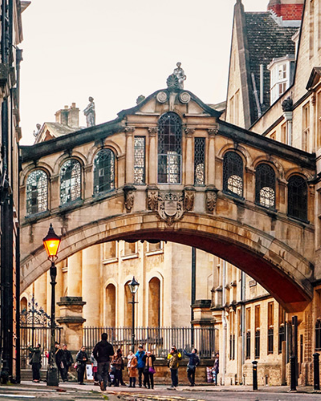 The Bridge of Sighs in Oxford — a covered stone bridge crossing New College Lane — photographed at dusk. An ornate arched bridge with mullioned windows connects two college buildings above a narrow street lit by a warm gas-style lamp. A small group of visitors gather below.