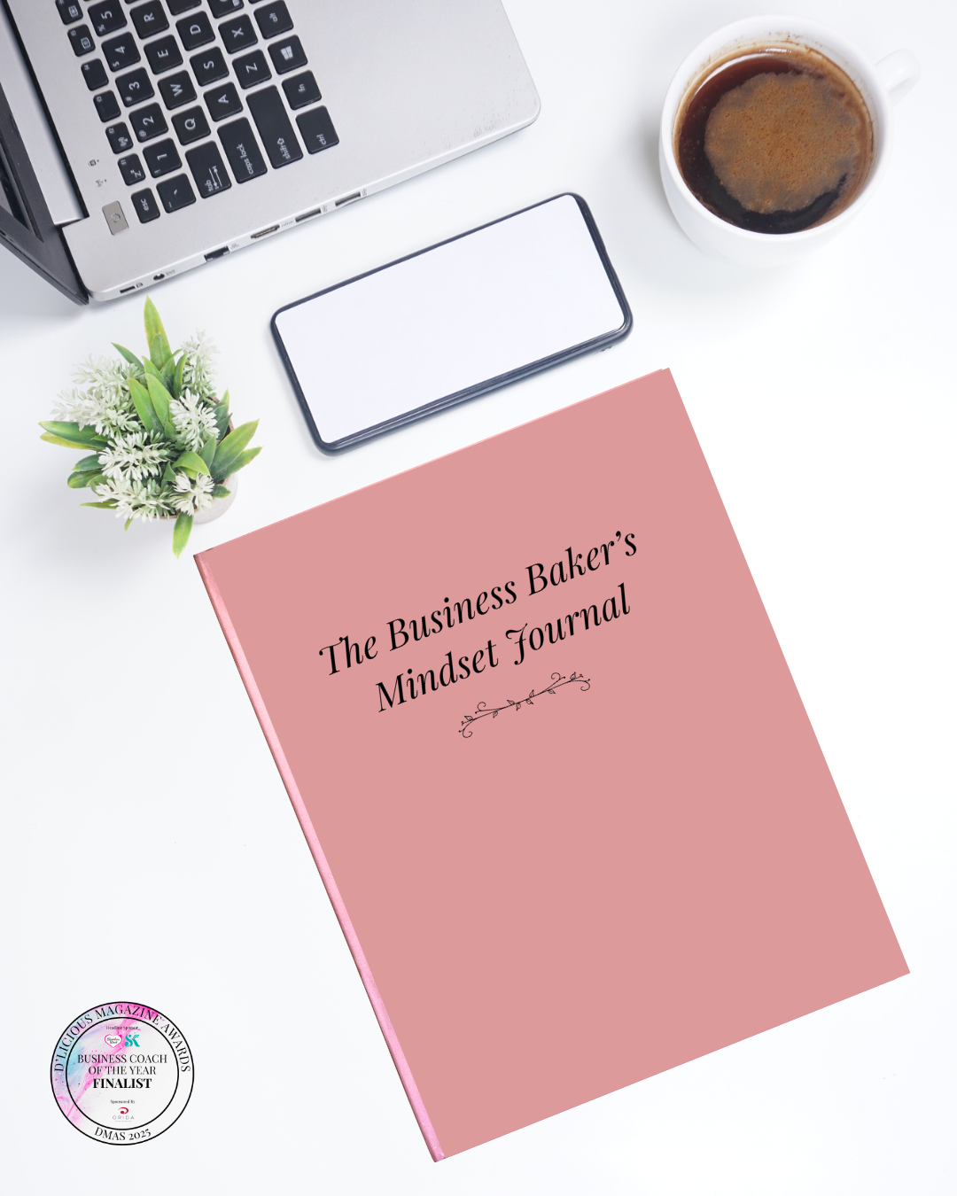 The Business Baker's Mindset Journal in rose pink, angled on a white desk alongside a laptop, smartphone, white plant pot, and cup of black coffee. D'Licious Magazine Awards Business Coach of the Year Finalist badge shown in the bottom left corner.<br />
